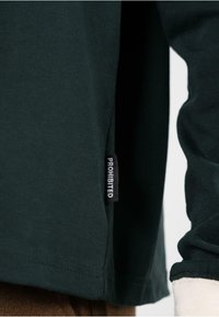 Camisa de manga larga negra con una textura suave. Cuenta con una etiqueta lateral que dice "PROHIBIDO" en blanco. El dobladillo está cosido con un acabado limpio.