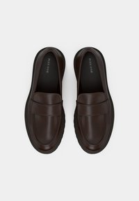 Καφέ δερμάτινα μοκασίνια με λεία φινίρισμα, ραμμένο σχέδιο και στρογγυλεμένη μύτη. Διαθέτουν ανυψωμένη λωρίδα πάνω από το πέλμα και υφή στη σόλα.