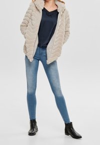 Beżowa kurtka z wzorem w zygzak z kapturem, zestawiona z czarną bluzką i jasnoniebieskimi rurkami, uzupełniona czarnymi botkami za kostkę.