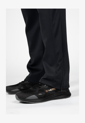 Zwarte sportbroek met een gestructureerd zijpaneel, gecombineerd met zwart-grijze sneakers met meshaccenten en oranje "GWEAR" branding.