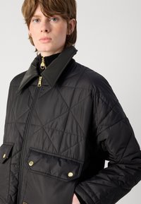 Casaco preto acolchoado com colarinho alto, detalhes em dourado, dois bolsos frontais e um padrão diagonal no tecido. Fecho com zíper.
