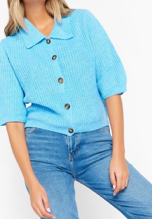 Frau trägt eine hellblaue Strickjacke mit braunen Knöpfen und kurzen Ärmeln, kombiniert mit blauen Jeans.