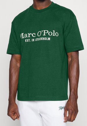Mężczyzna ubrany w ciemnozieloną koszulkę z białym napisem "Marc O’Polo EST. IN STOCKHOLM" oraz białe spodnie z zieloną haftowaną nazwą "Tiger".