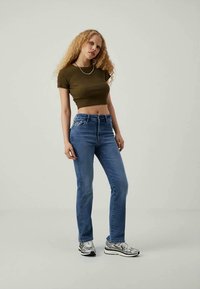 Mujer con cabello rizado, vistiendo una camiseta corta de oliva, jeans de tiro alto azules, zapatillas plateadas y un collar plateado, de pie contra un fondo liso.