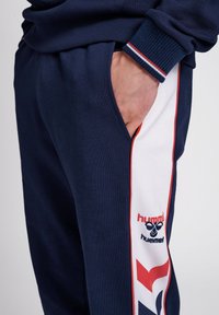 Marineblaue Sweatpants mit weißen Seitenstreifen und roten Akzenten, einem Logo und gerippten Bündchen. Hergestellt aus weichem Stoff mit einer lockeren Passform.