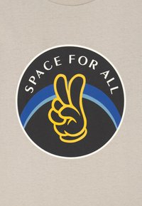 Keltainen ääriviiva käsistä, joka tekee rauhanmerkkiä ympyrän sisällä, jossa on teksti "KAIKILLE AURINKOA" ja sinisiä kaaria beige kankaan taustalla.