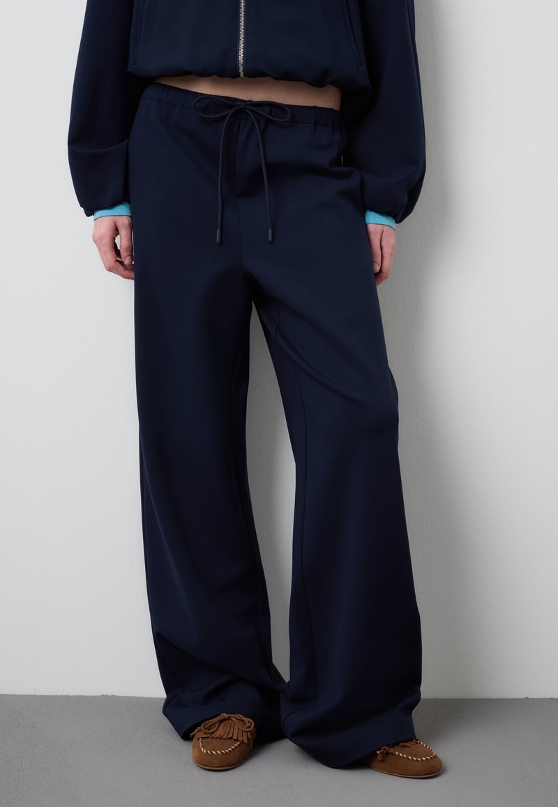 Persoon draagt marineblauwe wijde broek met trekkoord, een kort marineblauw jasje met turquoise manchetknopen en bruine mocassin-stijl schoenen tegen een witte muur.