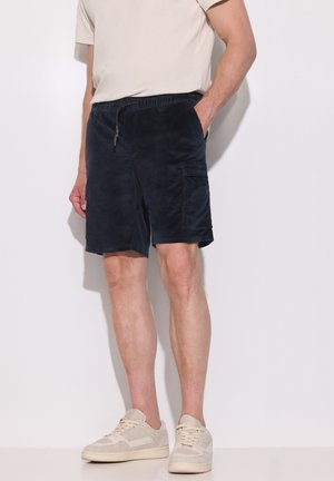 Homme portant un short en velours côtelé bleu marine avec poches avant et latérales, des baskets beiges et une chemise beige clair à manches courtes, debout contre un mur blanc.