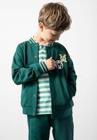 Zielona bomberka z białymi paskami na mankietach i kołnierzyku, ozdobiona haftowanymi naszywkami. Nosi się ją na białej koszulce polo w zielono-białe paski.