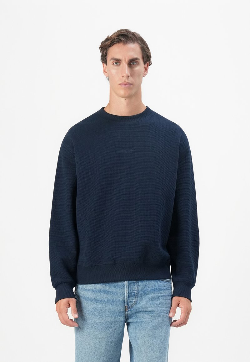 Marineblauwe textuurtrui met een ronde hals en geribde mouwen, gecombineerd met lichtblauwe jeans. Eenvoudig ontwerp, geen zichtbare logo's.