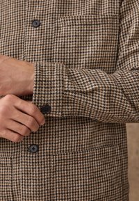 Giacca a scacchi marrone e beige con polsini abbottonati, dotata di tasca sul petto e tessuto testurizzato. Bottoni neri esaltano i dettagli di design.