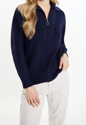 Pull en maille bleu marine avec un col zippé quart, présentant une texture côtelée et des manches longues, associé à un pantalon de couleur claire.
