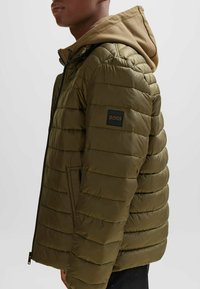 Chaqueta acolchada en verde oliva, con capucha color crema, costuras horizontales, bolsillos laterales y una etiqueta de logo negra en la manga izquierda.