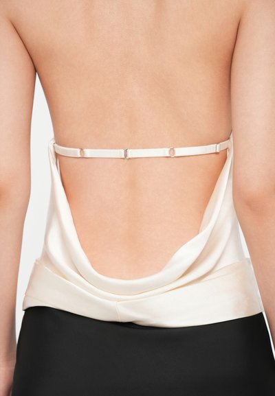 Πίσω όψη ενός κρεμ σατέν τοπ με χαμηλή λαιμόκοψη, που διαθέτει λεπτό, ρυθμιζόμενο λουρί και μαλακή, ριγέ υφή, συνδυασμένο με μαύρη φούστα.