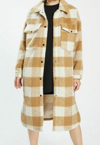 Lång rutig kappa i beige och vitt, tillverkad av mjukt, texturerat tyg. Har två framfickor och svarta knappstängningar.