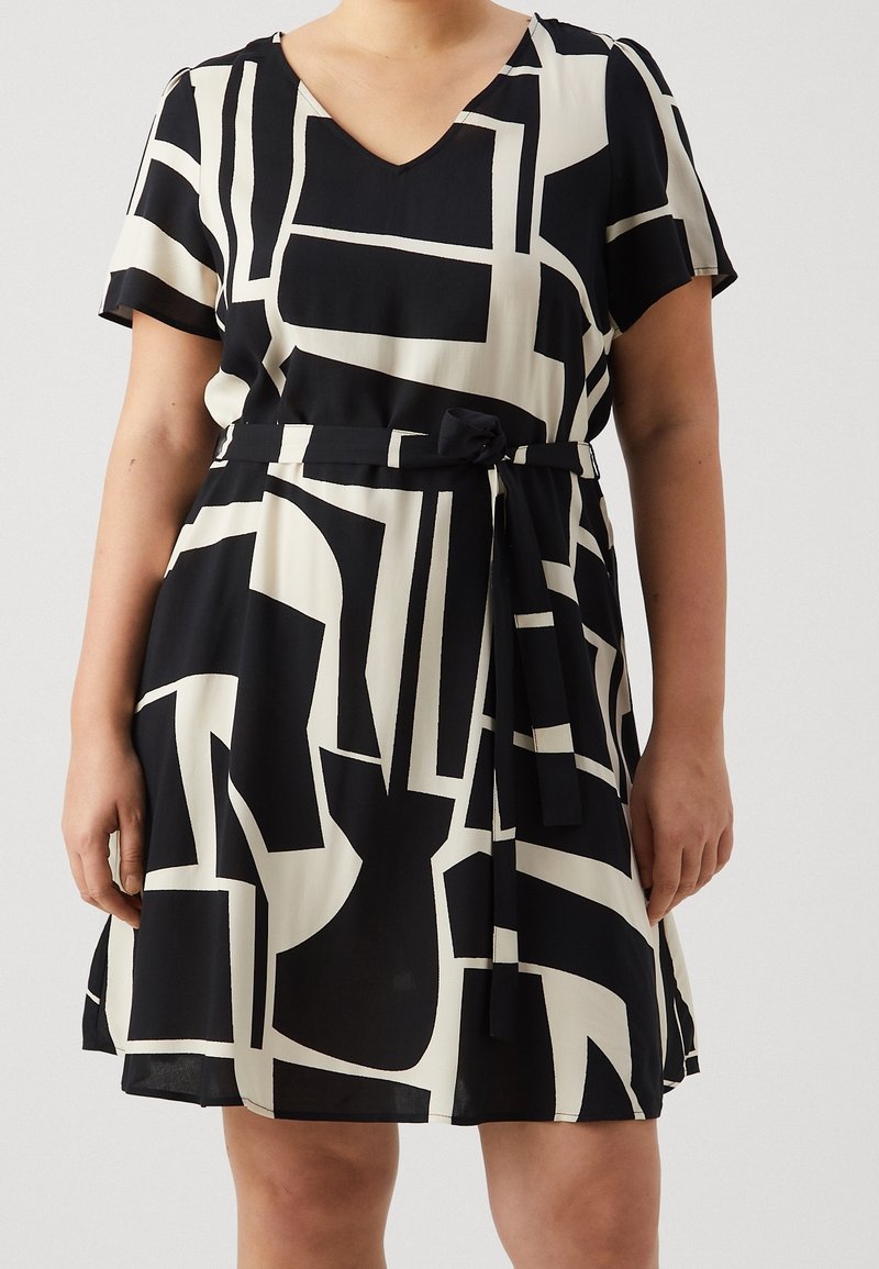 Robe à manches courtes, décolleté en V, présentant un motif géométrique abstrait noir et blanc, avec une taille cintrée et un détail de ceinture nouée. Tissu léger.
