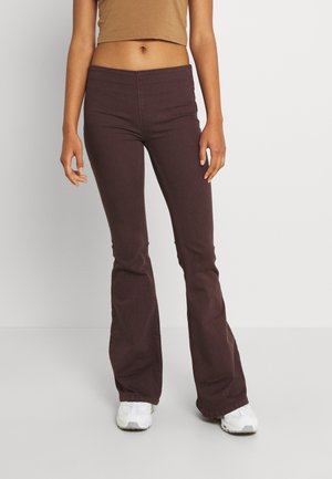 Weibliches Model trägt braune Schlaghosen, beigen kurzen Top und weiße Sneaker, steht vor einem einfarbigen hellgrauen Hintergrund.