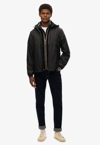 Zwarte lichtgewicht jas met een capuchon, ritssluitingen aan de voorkant en een contrasterende binnenvoering, gecombineerd met donkere denim jeans en crèmekleurige sneakers.