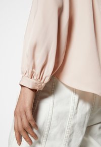 Bras gauche avec poignet de chemise à manches longues couleur pêche boutonné et main reposant à côté d’un pantalon blanc avec des coutures visibles.