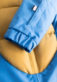 Polstret jakke med en blå yderflade med en glat tekstur, gul indvendig og elastiske manchetter. Inkluderer en synlig lynlås og syede detaljer.