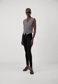 Szürke, szabott mellény V-nyakkivágással és hat gombbal, fekete leggingsszel és vastag fekete szandállal párosítva. Sima anyagtextúra, minimalista design.