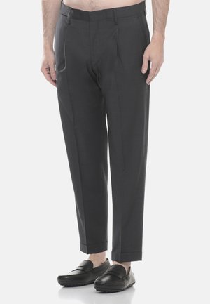 Uomo che indossa pantaloni neri su misura e mocassini neri, in piedi contro uno sfondo chiaro e uniforme.