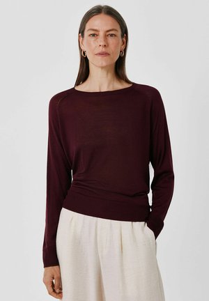 Vrouw met steil bruin haar draagt gouden oorringen, een donkerbordeauxrode longsleeve en lichtbeige broek tegen een effen achtergrond.