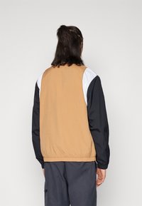 Kevyt takki väriblokki-kuvioinnilla; ruskea runko, mustat hihat ja valkoiset yksityiskohdat. Ominaisuuksina joustavat hihansuut ja rento istuvuus.