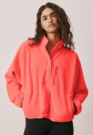 Femme aux cheveux ondulés portant une veste zippée corail vif avec des poches, debout les mains dans les poches devant un fond uni.