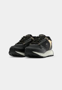 Baskets noires et beiges de la marque Replay avec des semelles blanches mouchetées, des lacets noirs avec des points jaunes, et un détail de logo sur le côté et la languette.