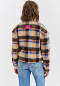 Vlněná kostkovaná bunda v béžové barvě s modrými, žlutými a burgundskými kostkami, s růžovým štítkem na zadní straně a knoflíkovými detaily na manžetách.