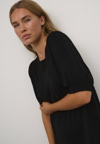 Robe tricot noire à la coupe décontractée, manches courtes bouffantes et poignets côtelés. Texture lisse avec un design simple et classique.