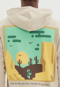 Felpa beige con un design grafico che rappresenta una scena desertica, sole, cactus e uccelli in tonalità di verde e giallo. Il testo aggiunge dettagli nella parte inferiore.