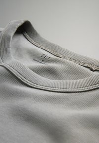 Helle graue Baumwoll-Sweatshirt mit geripptem Ausschnitt und einem Etikett mit der Aufschrift "SET". Geschmeidige Textur mit subtilen Steppdetails.