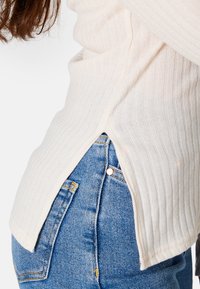 Rippentop in Creme mit seitlichen Schlitzen, kombiniert mit hellblauen Jeans aus Denim mit goldenen Akzenten und einer leicht ausgewaschenen Textur.