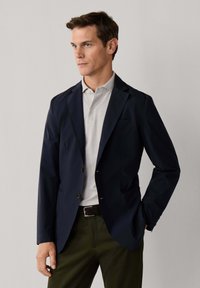 Muž ve tmavě modrém saku přes světle šedé polo tričko, s tmavě olivovými kalhotami a černým opaskem, stojící před jednoduchým pozadím.