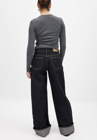 Jeans de denim oscuro de pierna ancha con dobladillo enrollado y costuras doradas. Presenta una etiqueta de cuero con "NA-KD" en la cintura trasera.