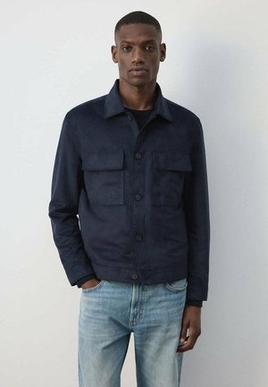 Hombre con una chaqueta azul marino oscuro abotonada con dos bolsillos en el pecho y vaqueros azul claro, de pie frente a un fondo claro y liso.