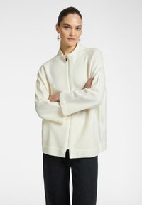 Donna con i capelli scuri, indossa un maglione bianco con zip e pantaloni neri, in piedi con le braccia incrociate, porta orecchini penzolanti a forma di stella.