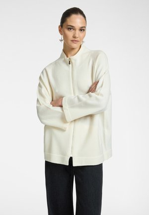 Donna con i capelli scuri, indossa un maglione bianco con zip e pantaloni neri, in piedi con le braccia incrociate, porta orecchini penzolanti a forma di stella.