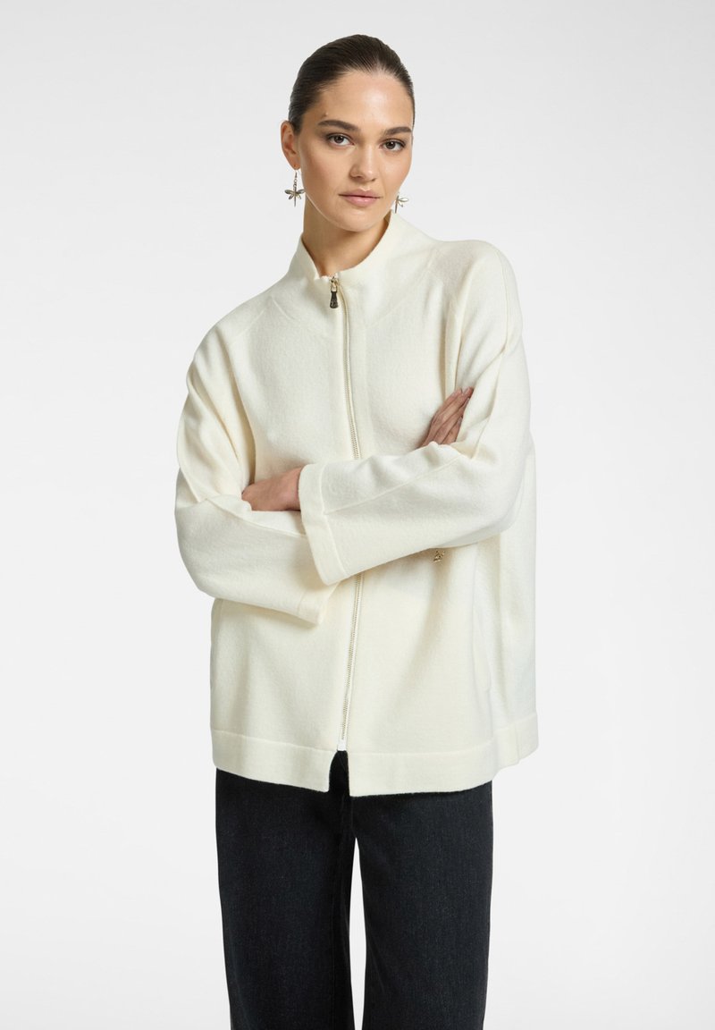 Donna con i capelli scuri, indossa un maglione bianco con zip e pantaloni neri, in piedi con le braccia incrociate, porta orecchini penzolanti a forma di stella.