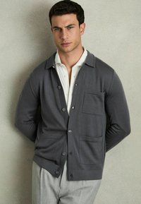Grå cardigan med krave-design, frontknapper og brystlomme, lavet af glat materiale, parret med en hvid skjorte og lyse bukser.