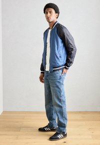 Blau-schwarze Bomberjacke mit gestreiften Bündchen, getragen über einem weißen Hemd, kombiniert mit hellblauen Jeans und schwarzen Sneakers.