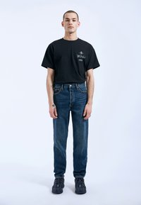 Camiseta de algodón negra con estampado gráfico en el pecho, combinada con pantalones vaqueros azules holgados y zapatillas negras. Atuendo simple y casual.