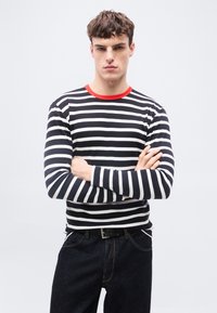 Νεαρός άνδρας με σταυρωμένα χέρια, φοράει μακρυμάνικη μπλούζα με ασπρόμαυρες ρίγες και κόκκινο γιακά, καθώς και σκούρα τζιν, μπροστά σε απλό φωτεινό φόντο.