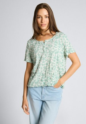 Junge Frau mit braunen Haaren, die eine hellgrüne Bluse mit Blumenmuster und kurzen Ärmeln sowie hellblaue Jeans trägt, steht mit einer Hand in der Tasche.