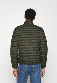 Donkergroen gewatteerde jas met een quilted ontwerp. Heeft een hoge kraag en elastische manchetten. Gemaakt van lichtgewicht materiaal, gecombineerd met blauwe denim jeans.