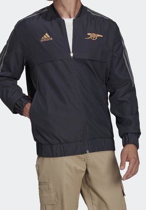 Homme portant une veste zippée noire Adidas avec logo Arsenal doré et pantalon cargo beige, debout avec une main dans la poche.