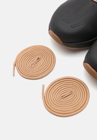 Mustat urheilukengät, joissa on teksturoitu pinta, ruskeat kumikoristeet ja kaksi vaaleanbeigeä kengännauhaa, jotka on asetettu kierteiksi.