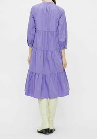 Vestido midi morado, con capas, y mangas 3/4, de tela ligera, escote redondeado y silueta fluida, combinado con botas blancas hasta la rodilla.